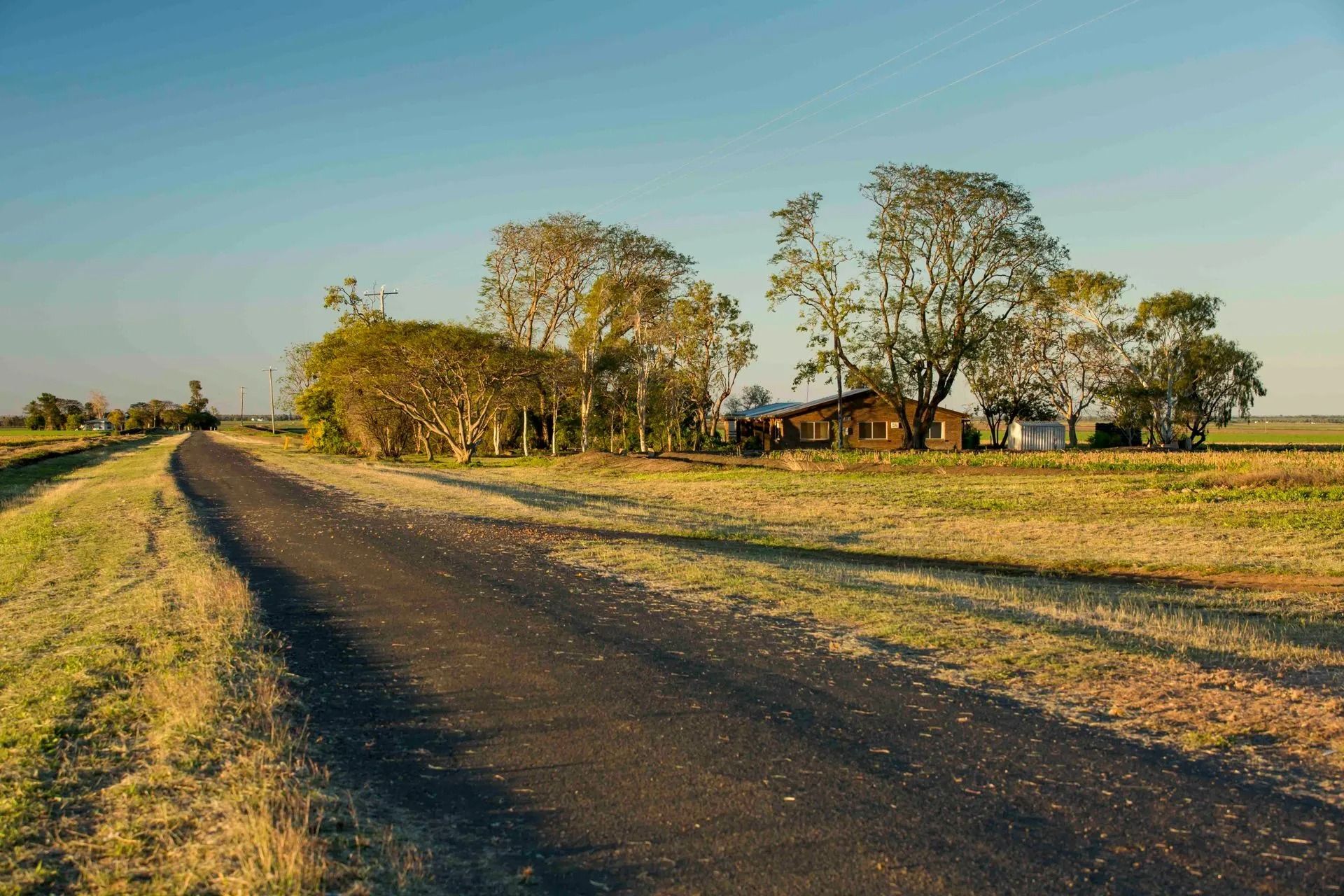 A rural paved road winds through a dry grassy landscape toward a house nestled among trees under a clear blue sky.