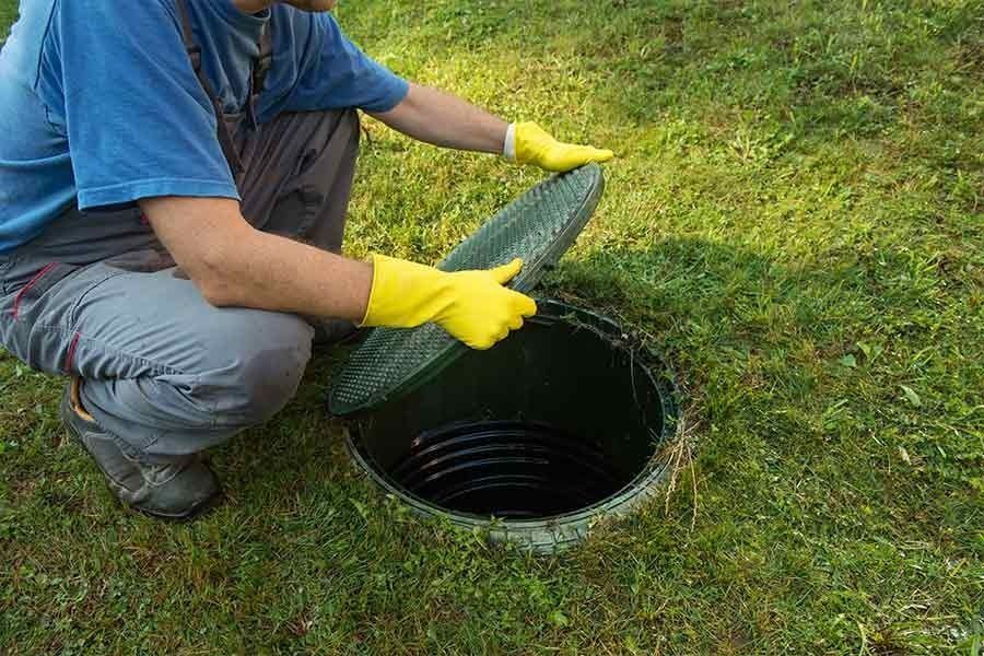 A person wearing yellow rubber gloves kneeling on the grass and lifting the lid off a circular septic tank access port.