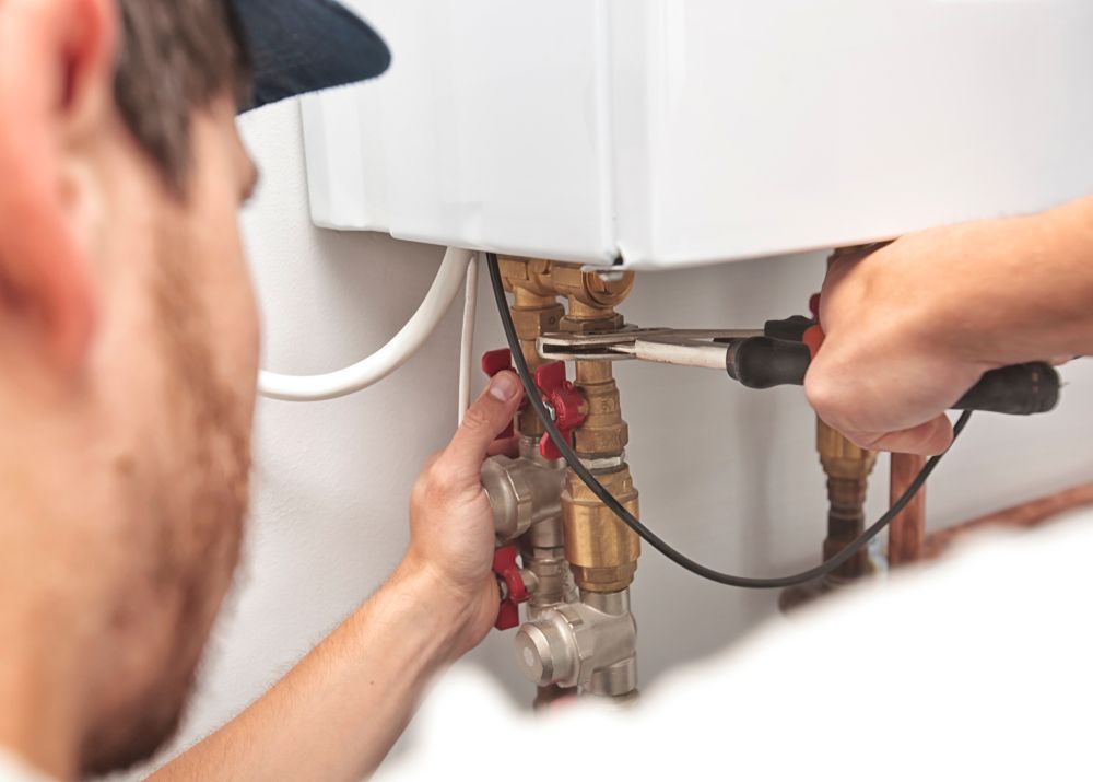 A person using pliers to adjust brass valves on the underside of a white wall-mounted boiler.