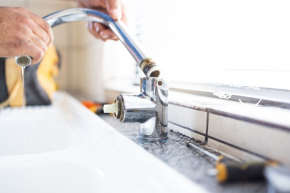 A person repairs a kitchen faucet, holding the disconnected spout above the base on a stone countertop with tools nearby.