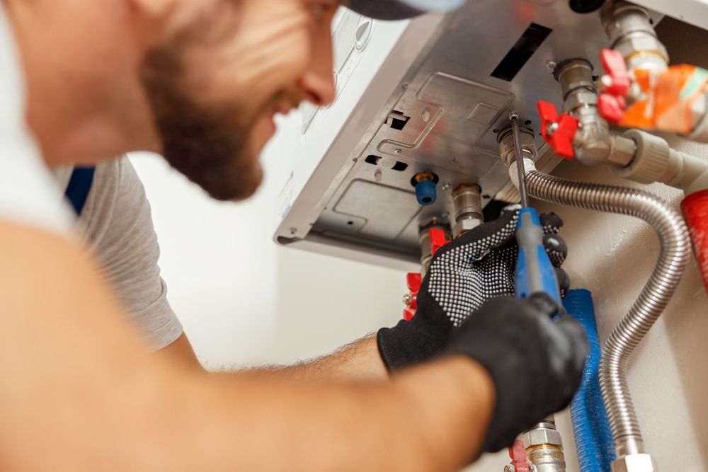 A technician wearing work gloves uses a screwdriver to repair the plumbing connections on a wall-mounted boiler unit.