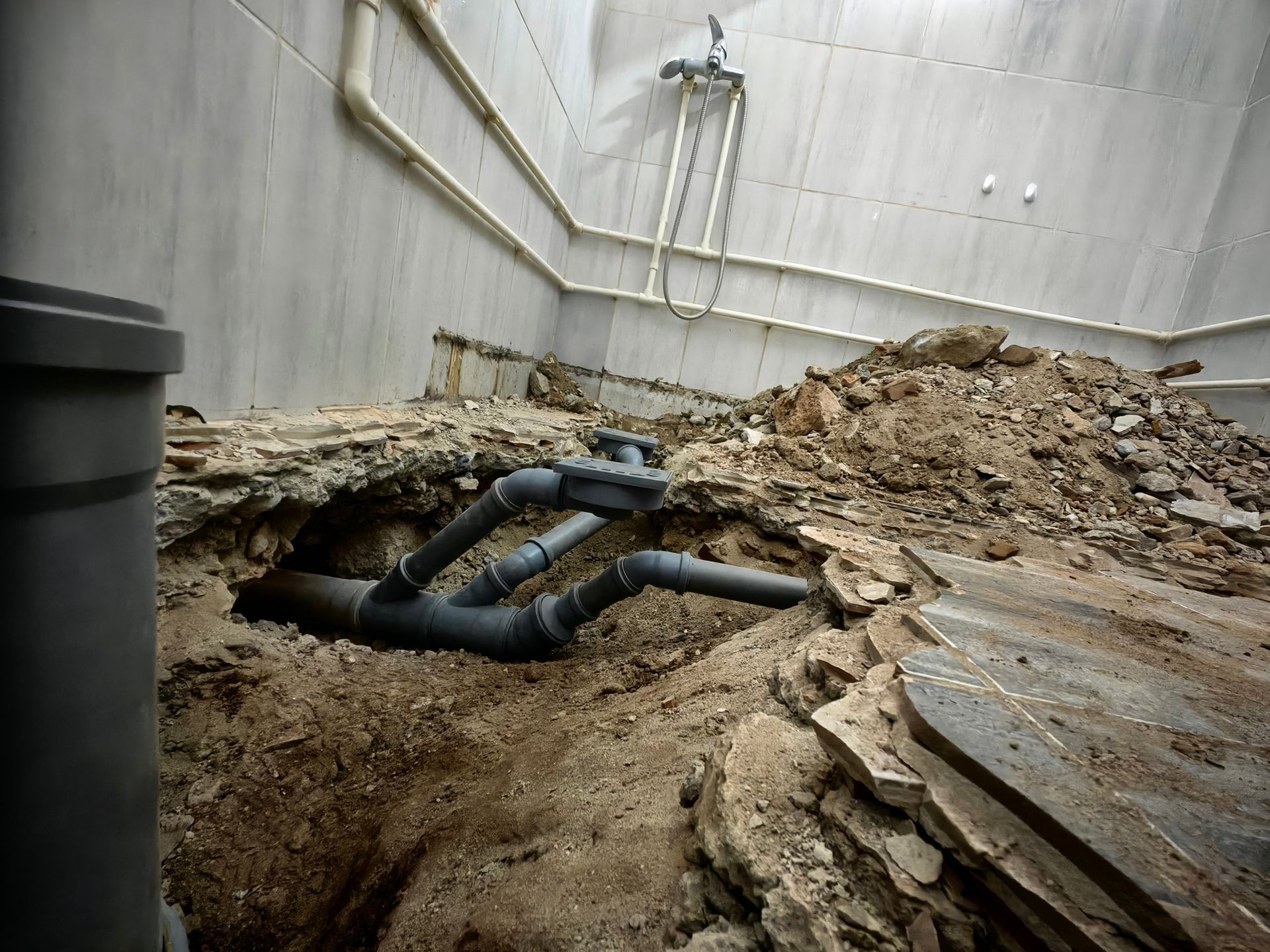 An excavated room floor showing exposed grey plumbing pipes in a dirt trench, with white tile walls and a shower head.