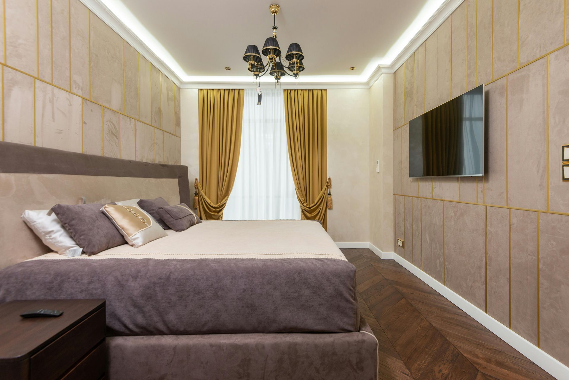 Bedroom with upholstered bed, gold curtains, TV, and wood floor.