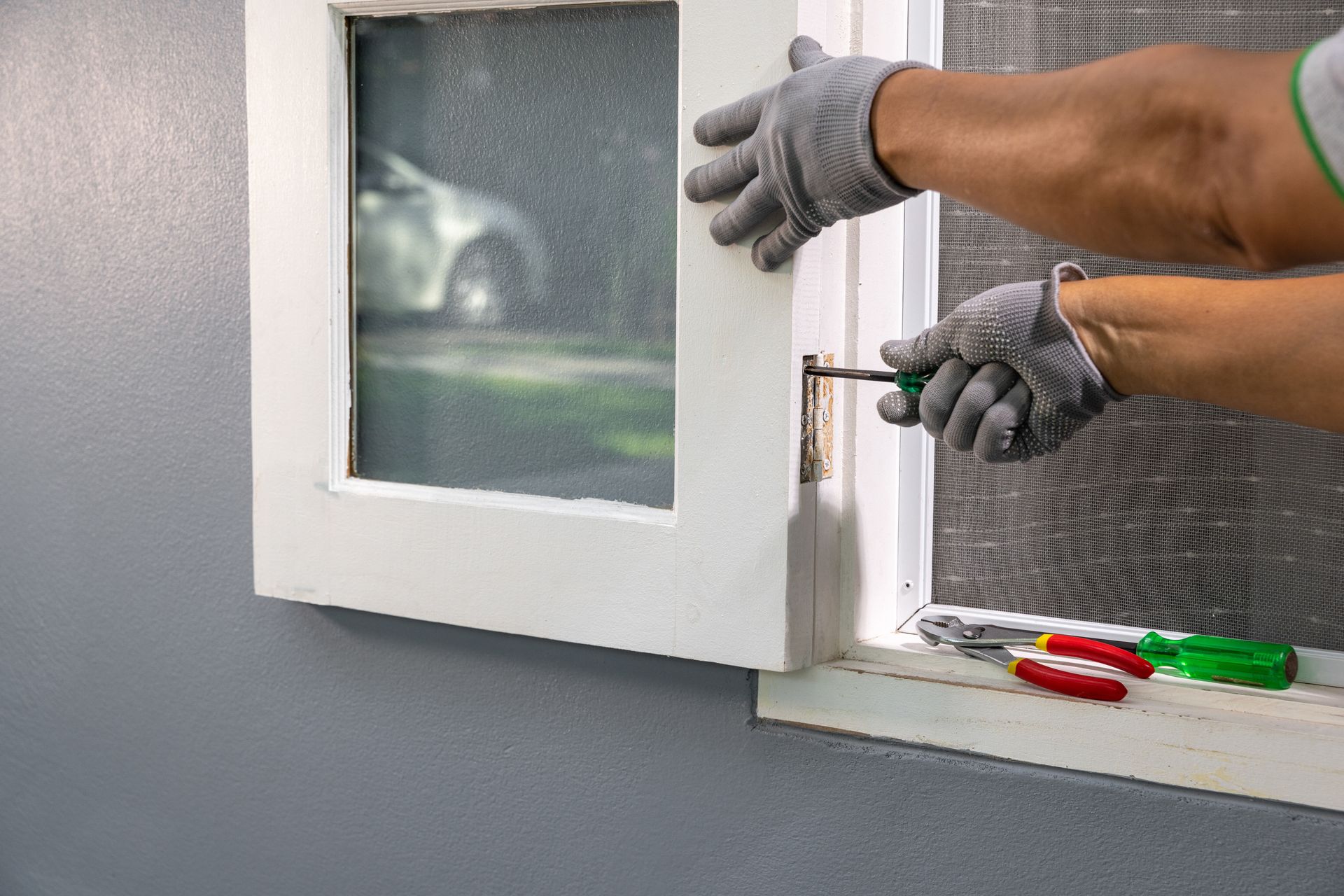 Person with gloves using a screwdriver to fix a white window frame on a gray wall.