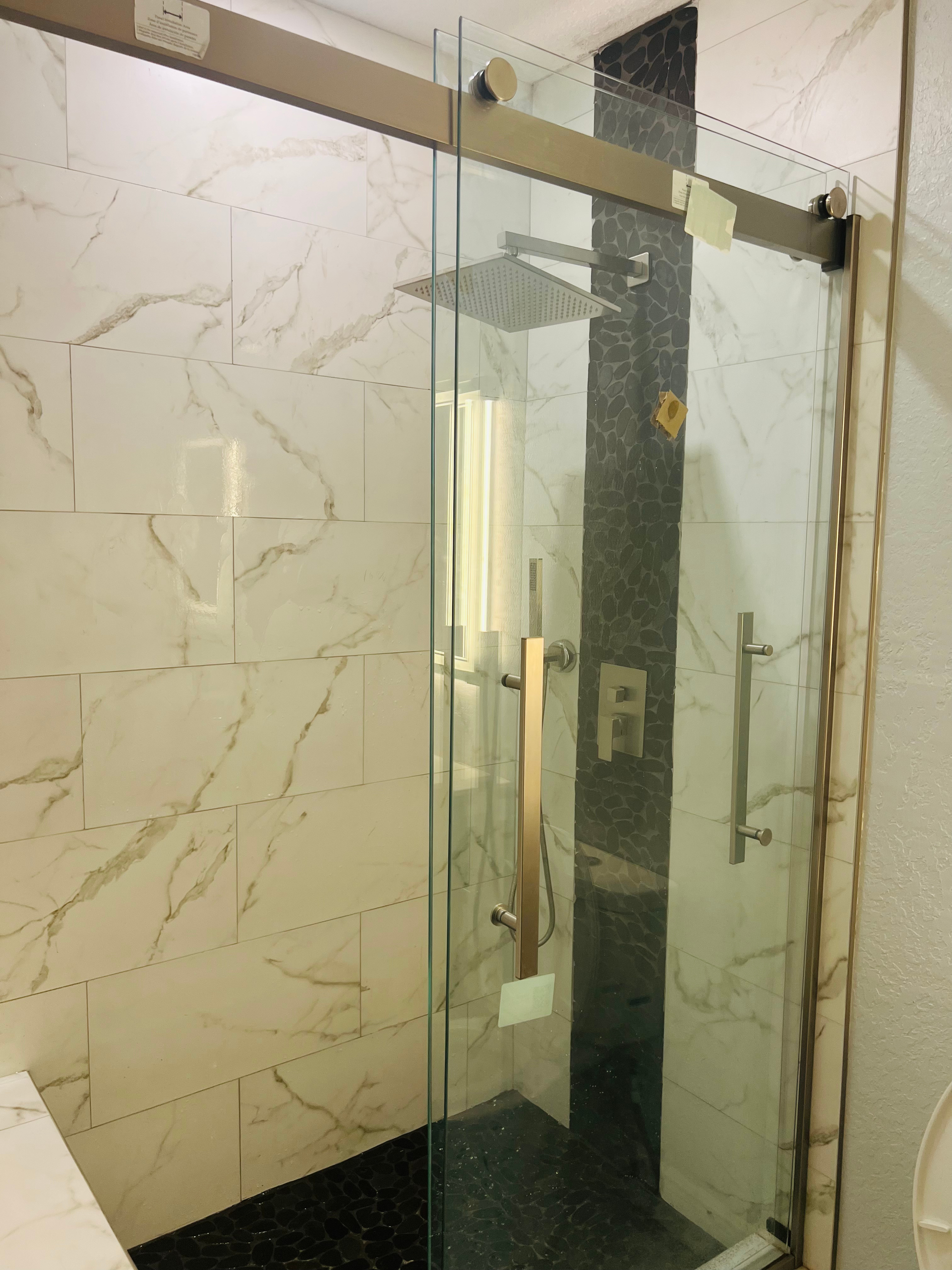 Glass shower enclosure with sliding door, gold fixtures, marble tile wall.
