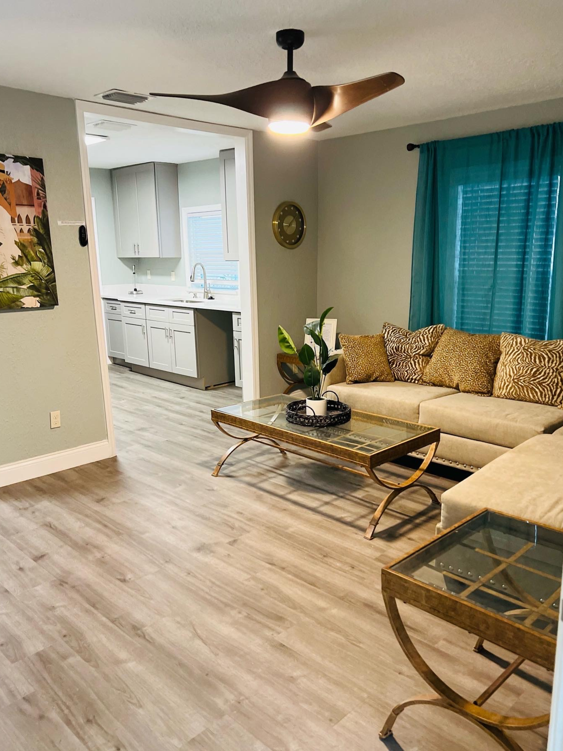 Living room with light gray walls, open to kitchen, tan couch with animal print pillows, turquoise curtains.