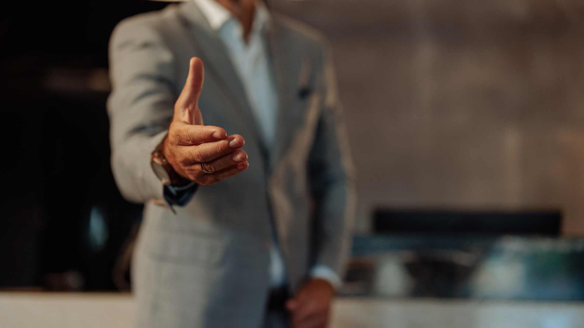 Man in a gray suit extends hand for a handshake, arm outstretched, indoors.