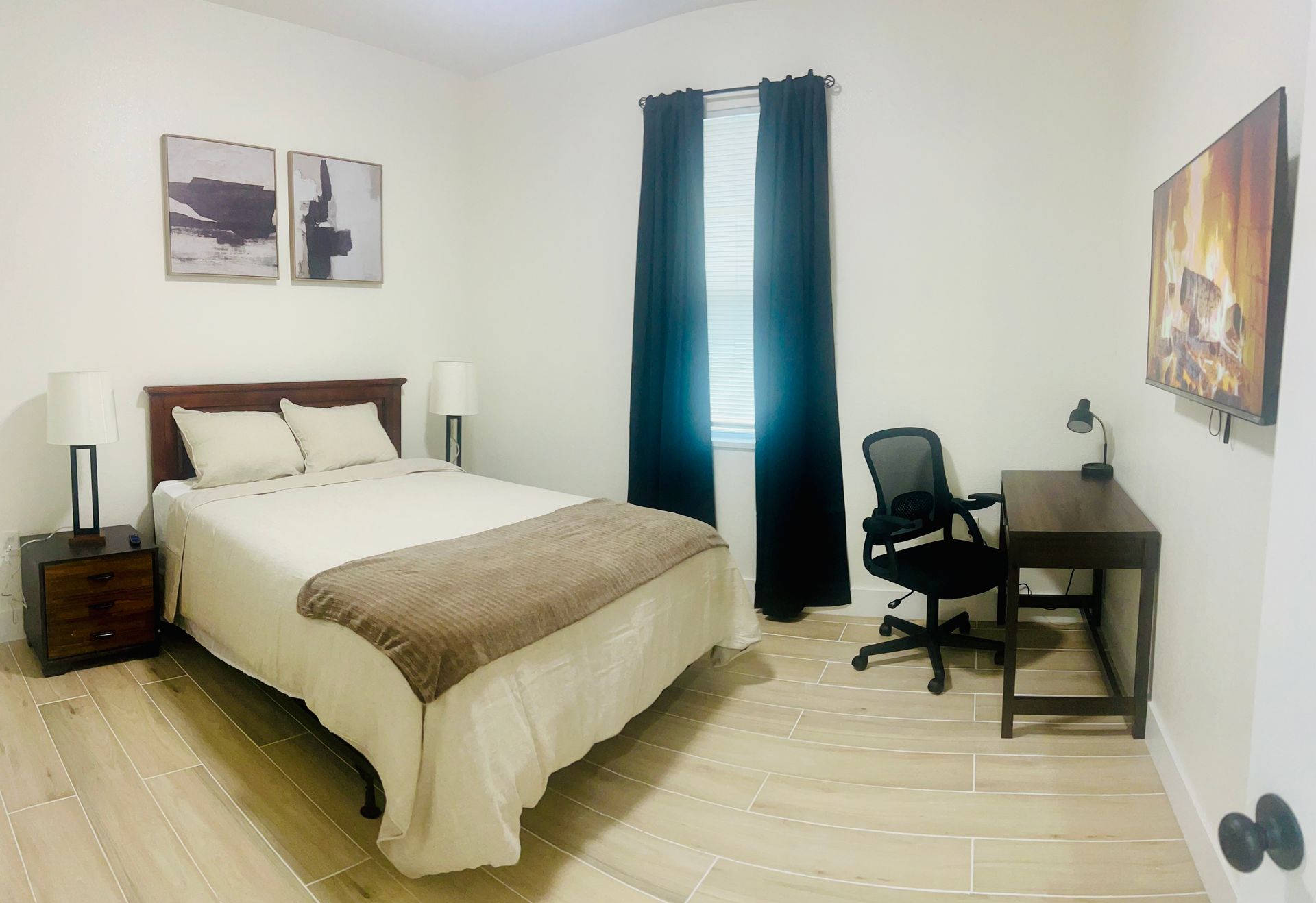 Bedroom with bed, desk, artwork, and window with black curtains.