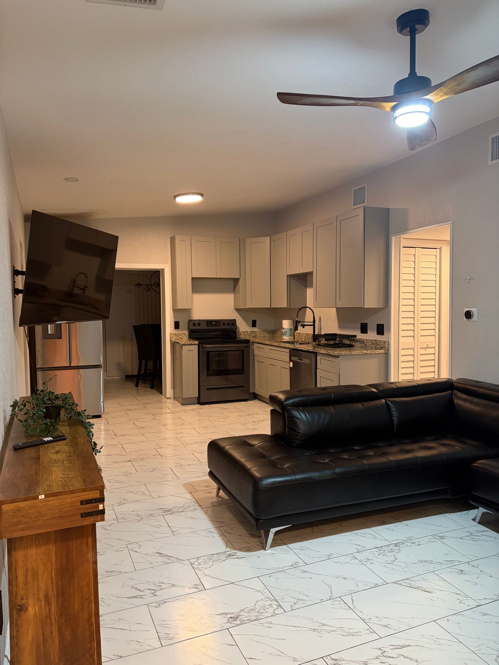 Living room with black leather sectional sofa, open to a kitchen with gray cabinets and a mounted TV.
