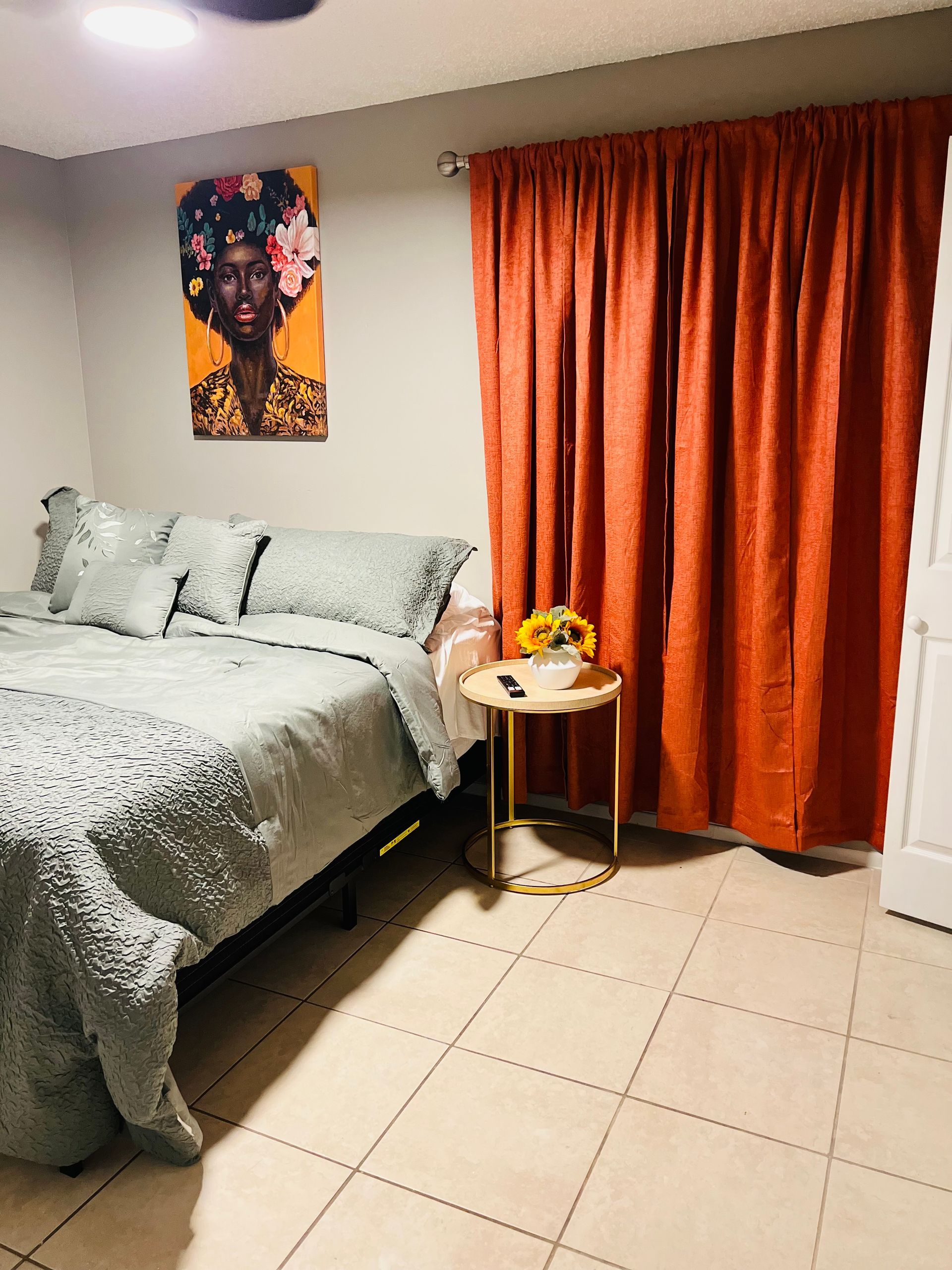 Bedroom with bed, art, orange curtains, and side table with sunflowers.