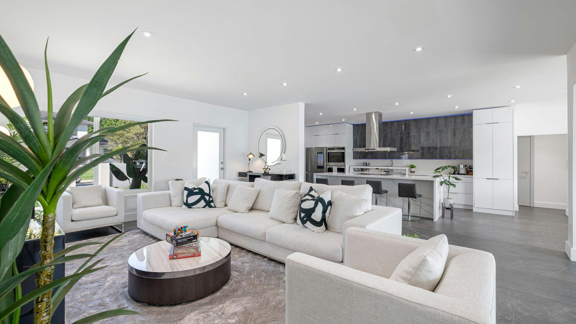 Bright modern living room with white sofas, round coffee table, large plant, and open kitchen in the background