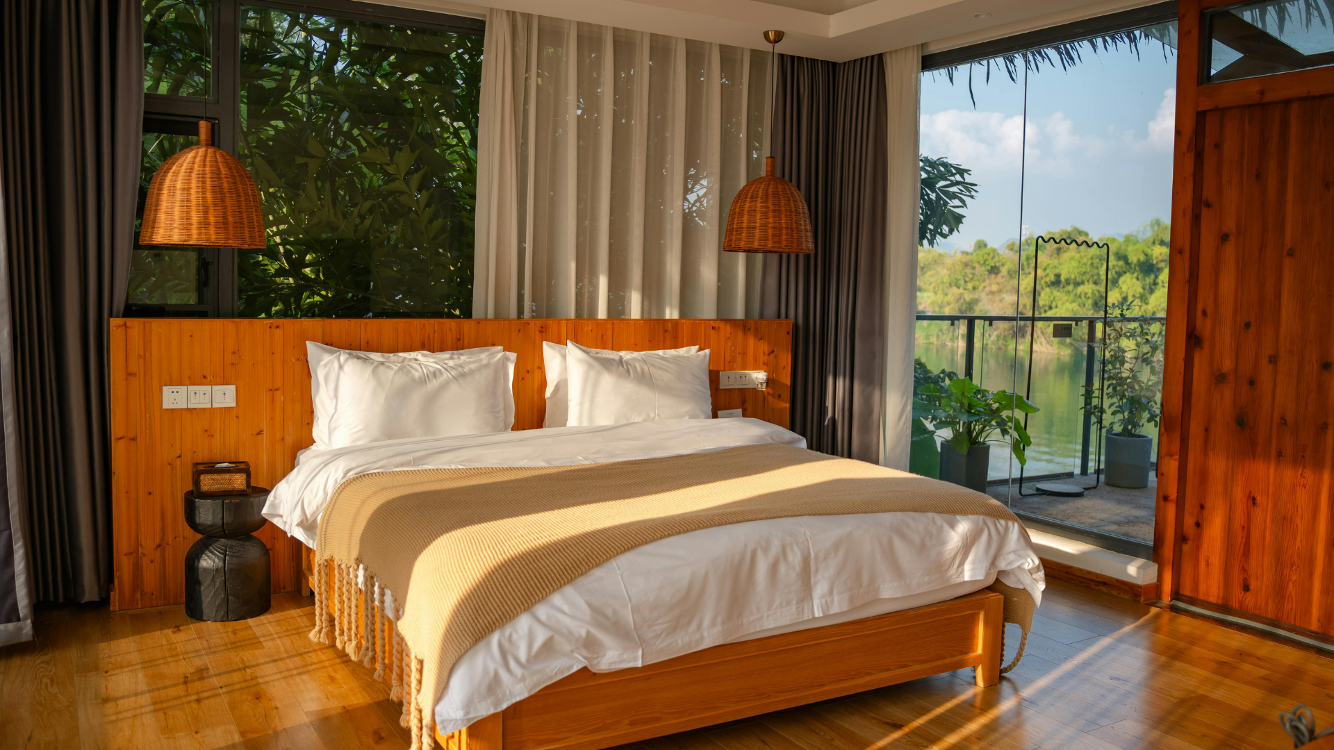 Modern bedroom with wooden bed, white bedding, pendant lamps, and a large window overlooking greenery
