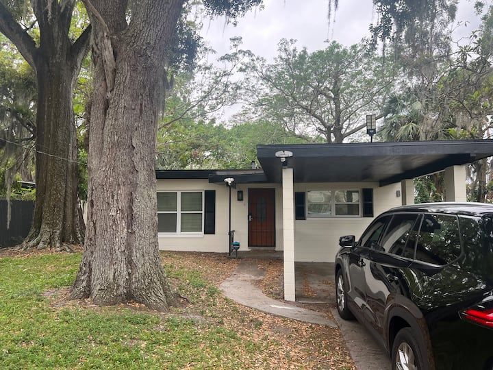 Small, beige house with black accents, covered carport, and a black car parked in the driveway.