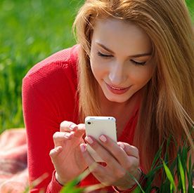 Woman with long blonde hair, wearing a red shirt, looking at a white phone while lying in green grass.