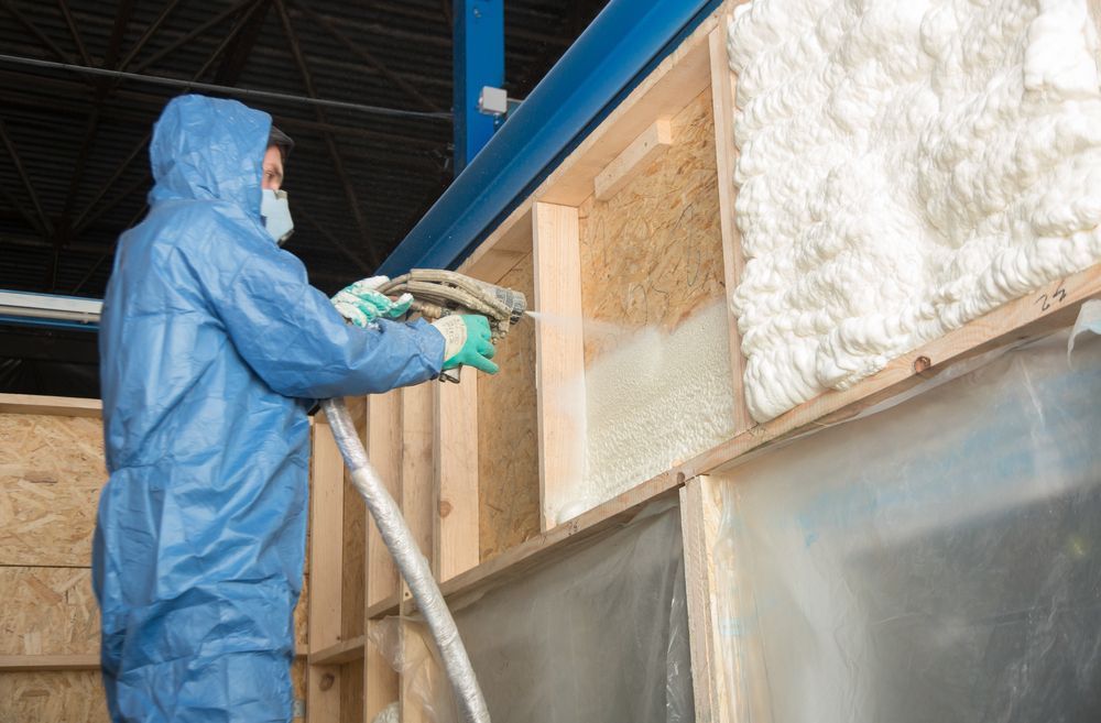 Person in protective suit sprays foam insulation onto a wooden wall indoors.