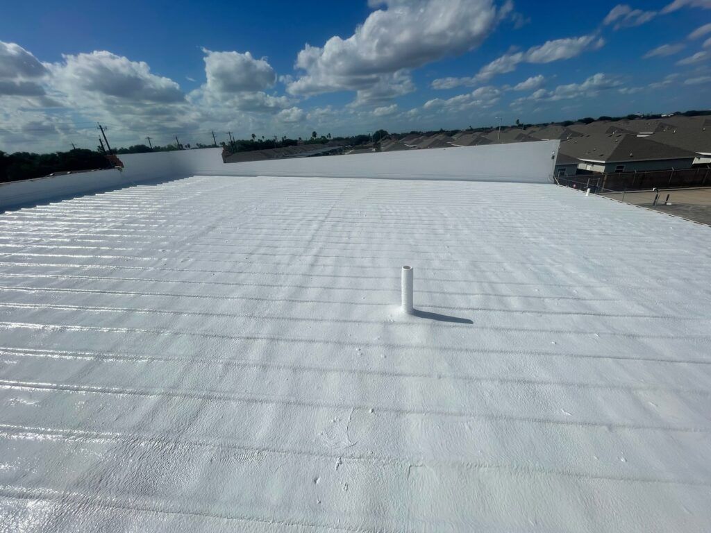 White coated flat roof under a bright blue sky with clouds, a vent pipe, and a building in the background.