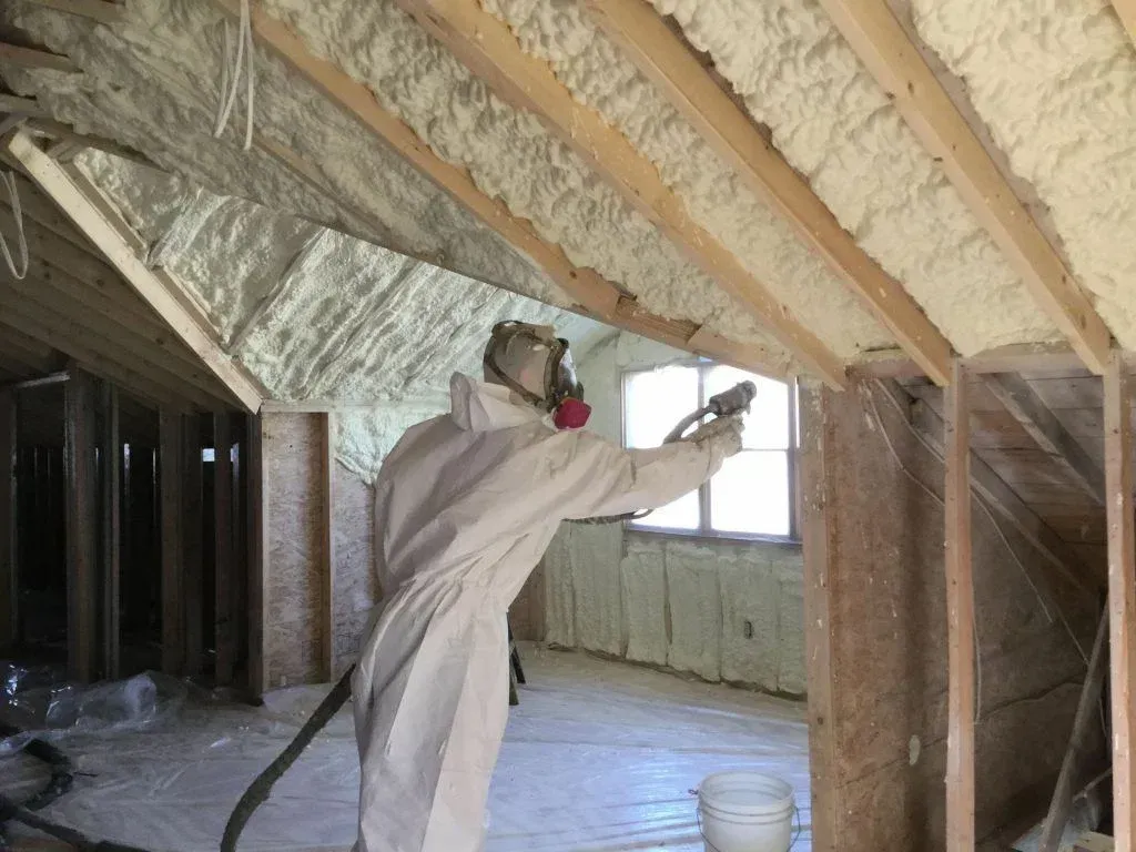Person in protective suit spraying foam insulation in an attic with wooden beams.