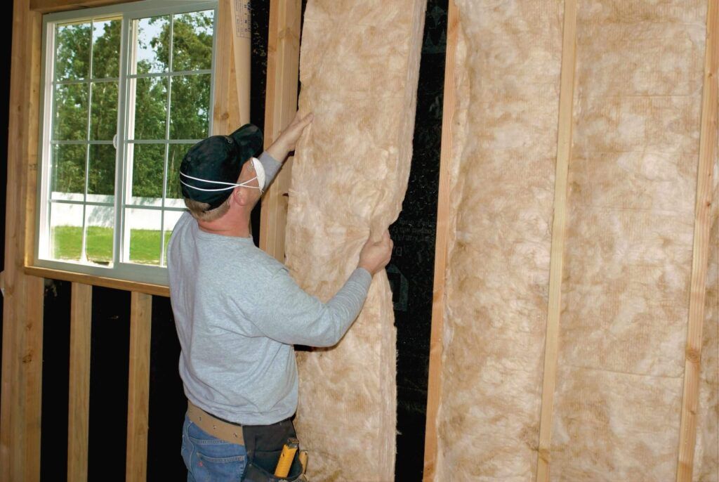 Man installing insulation in a wall, wearing a mask and safety glasses. Window in background.
