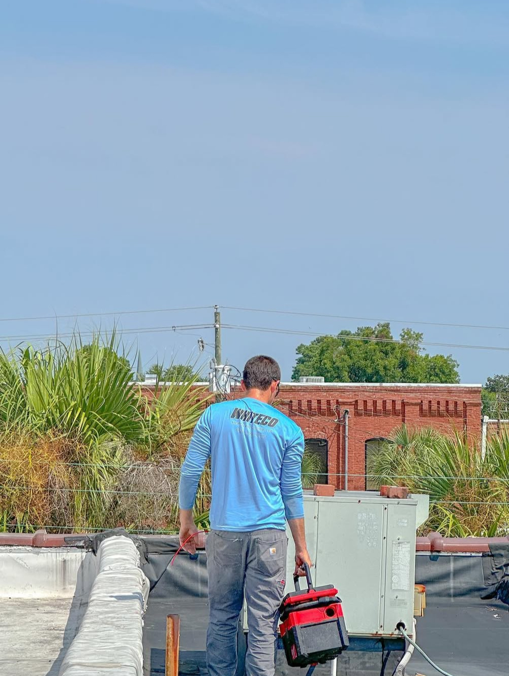 A man is standing on top of a roof holding a tool bag.