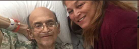 A woman smiles next to a bald man in a hospital bed, both looking at the camera.
