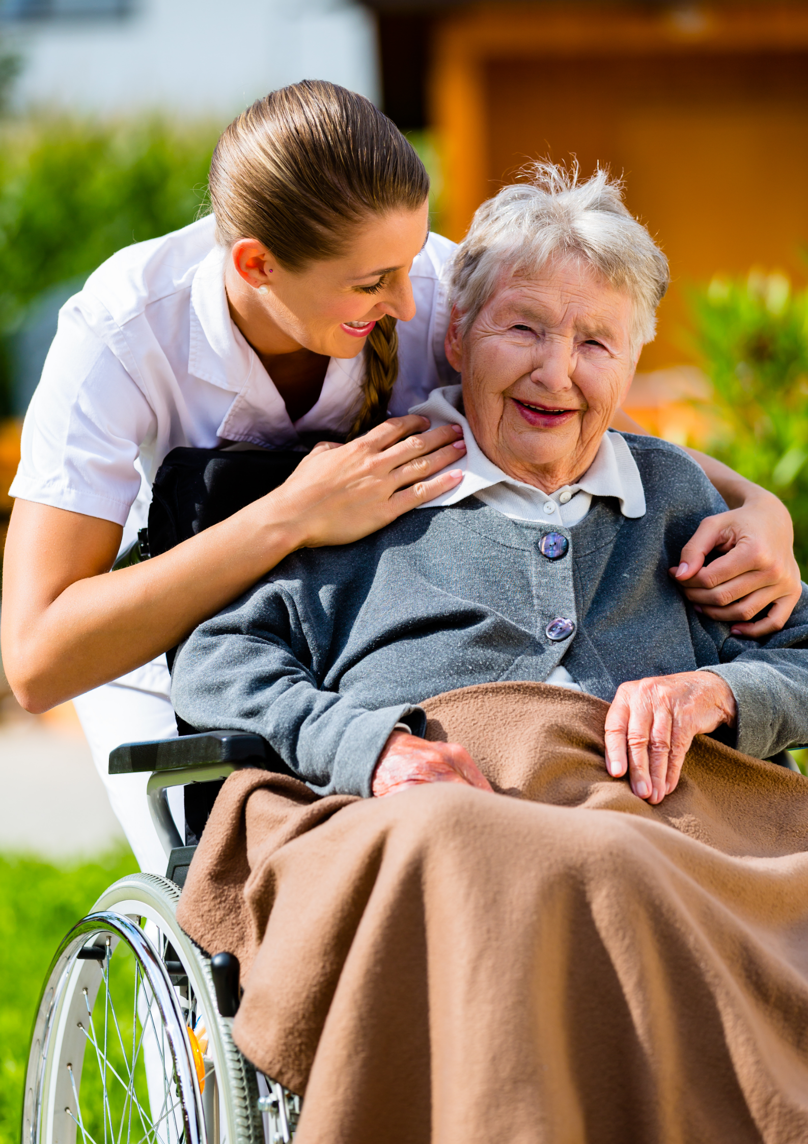 Woman in white scrubs assists an elderly woman in a wheelchair outdoors, smiling.