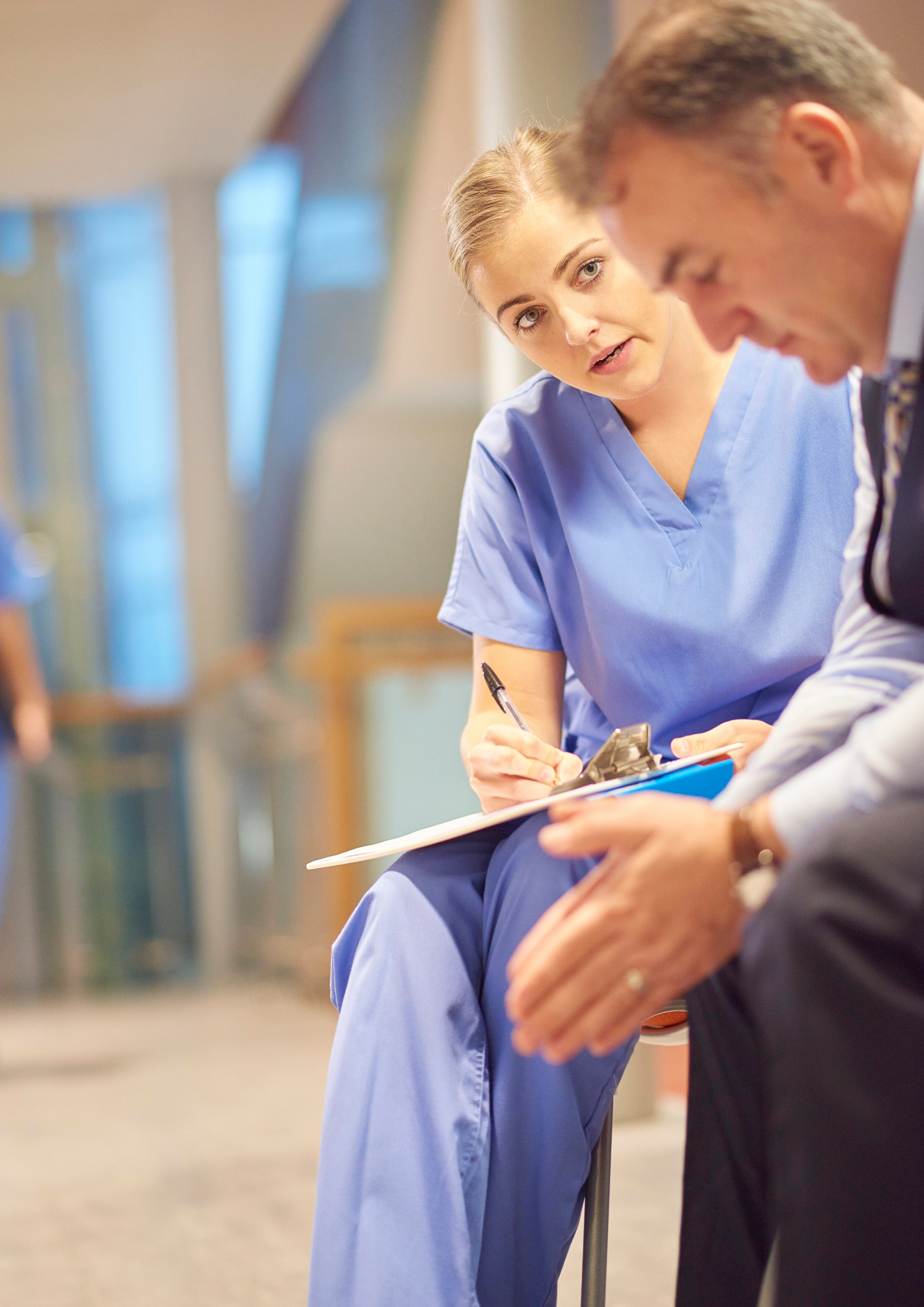 Nurse in blue scrubs takes notes while speaking with a man in a light-filled room.