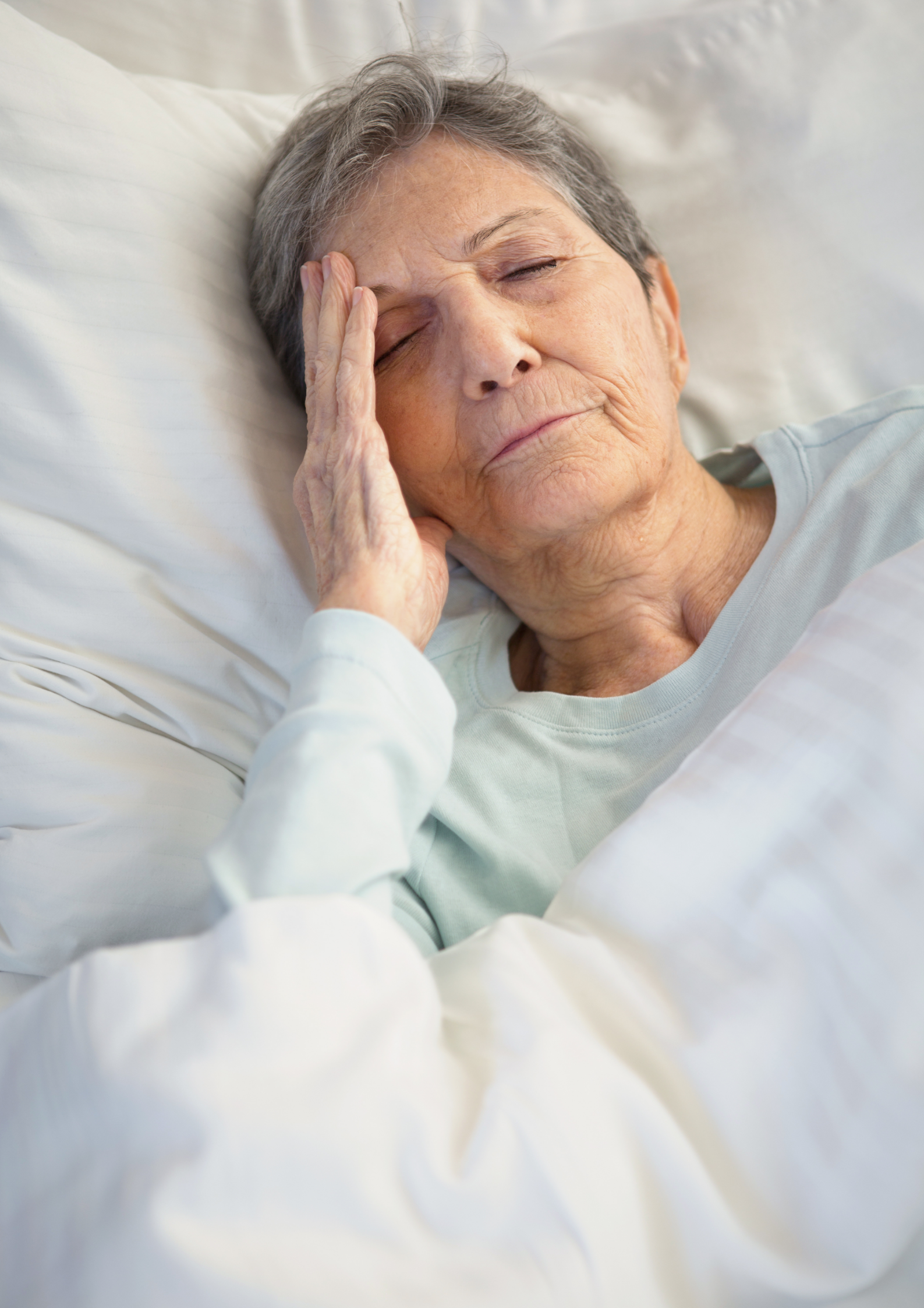 Elderly woman in bed, eyes closed, hand on forehead; possibly unwell.