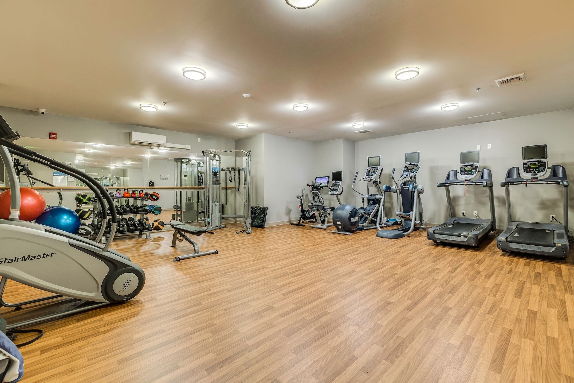 A well-lit gym with various exercise machines, weights, and a mirrored wall. Wooden floor.
