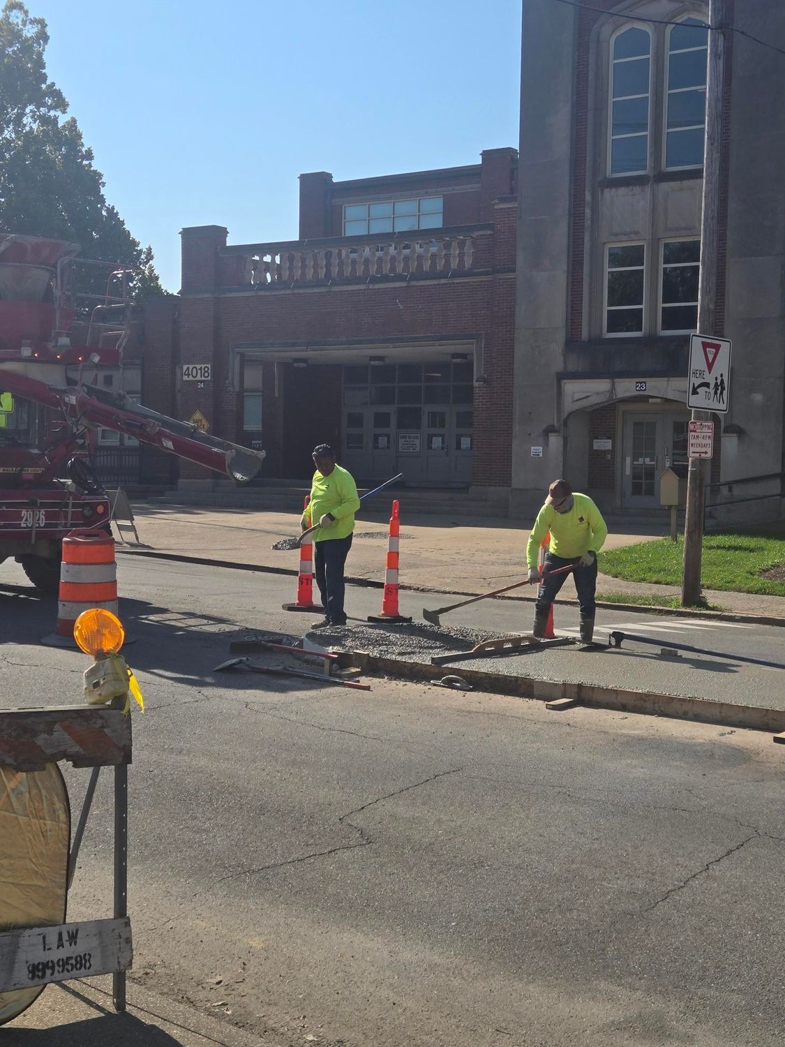 Workers in safety vests repair road near a brick building. Orange cones and equipment are present. Sunny day.