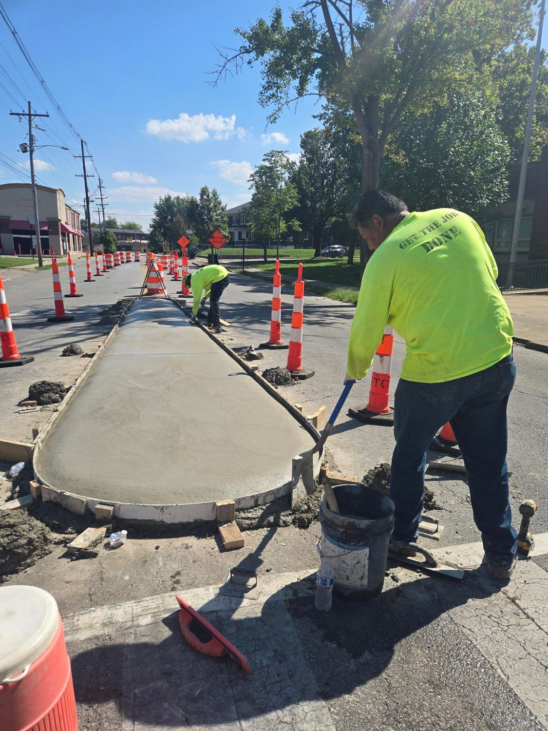Construction workers pouring concrete on a street with traffic cones, on a sunny day.