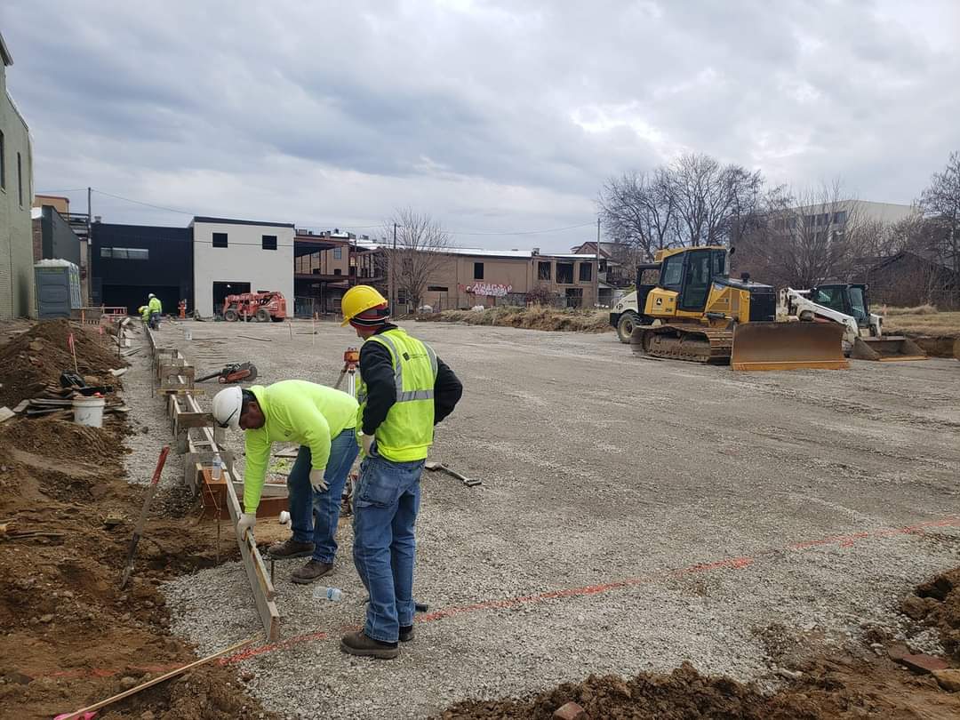 Construction workers on a gravel site, wearing hard hats and vests, with machinery and buildings in the background.