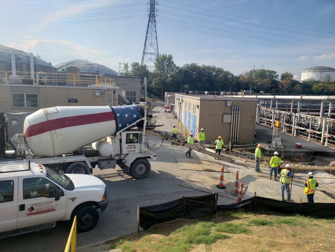 Cement truck with American flag design pouring concrete at a construction site; workers in yellow vests.