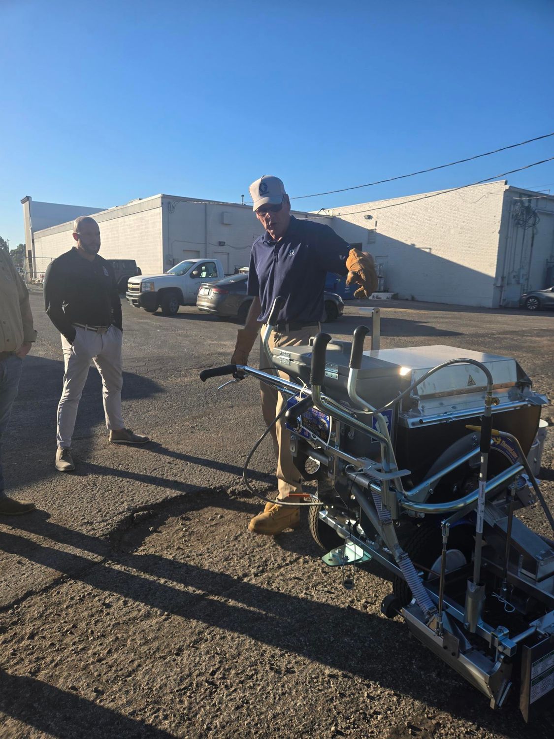 Man operating line painting machine on asphalt, another man watching in front of a white building.