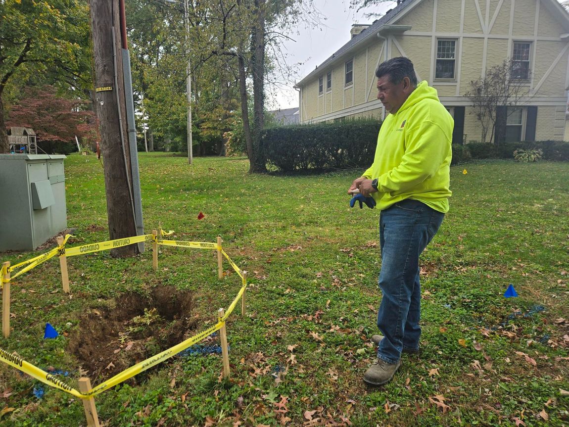 Man in neon yellow jacket beside a dirt excavation surrounded by yellow tape and markers on a lawn.