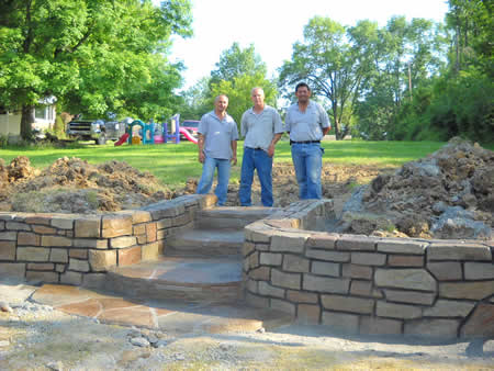 Three men standing on stone steps, newly constructed wall. Green trees and lawn in the background.
