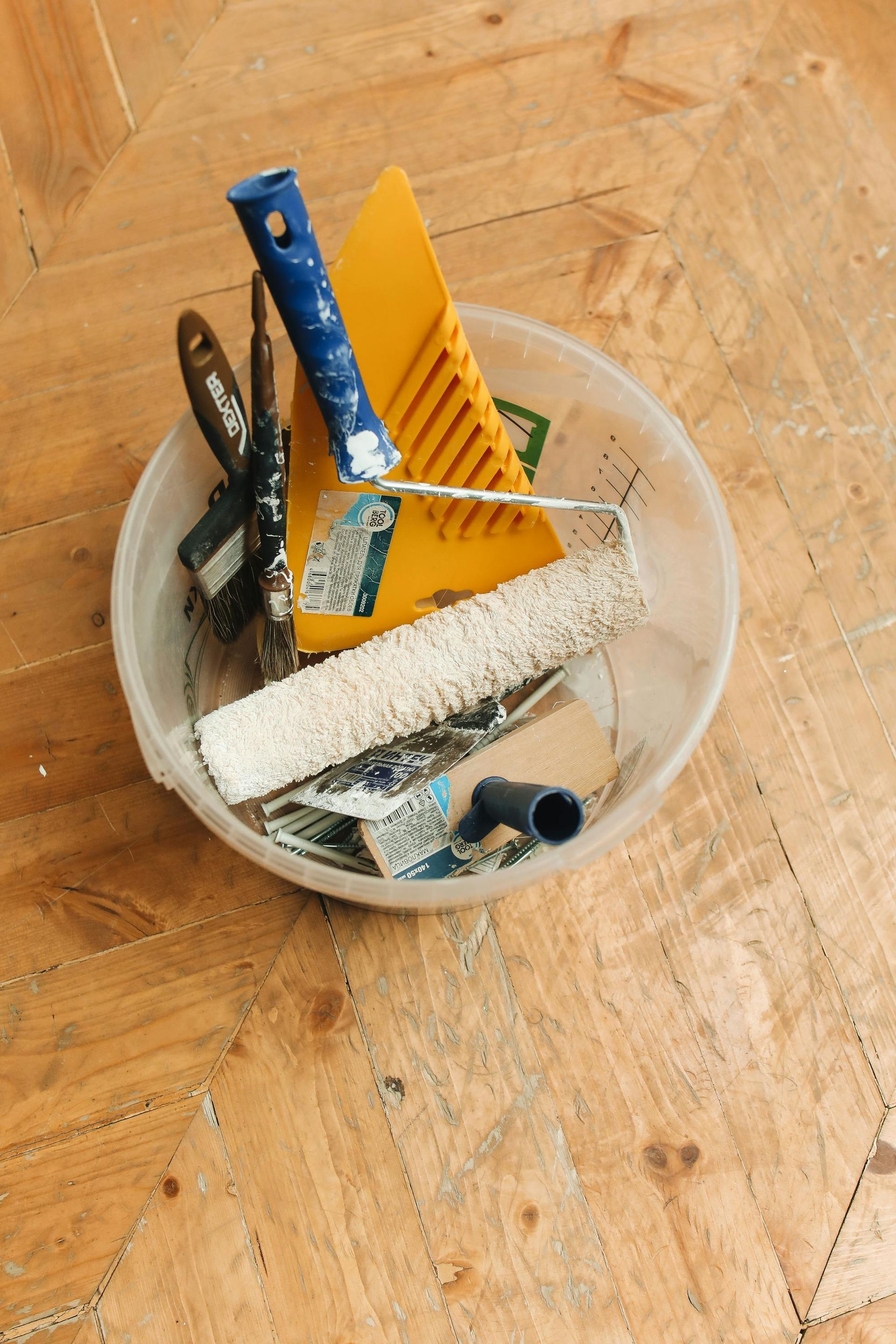 A bowl filled with paint rollers , spatulas , and other tools on a wooden floor.