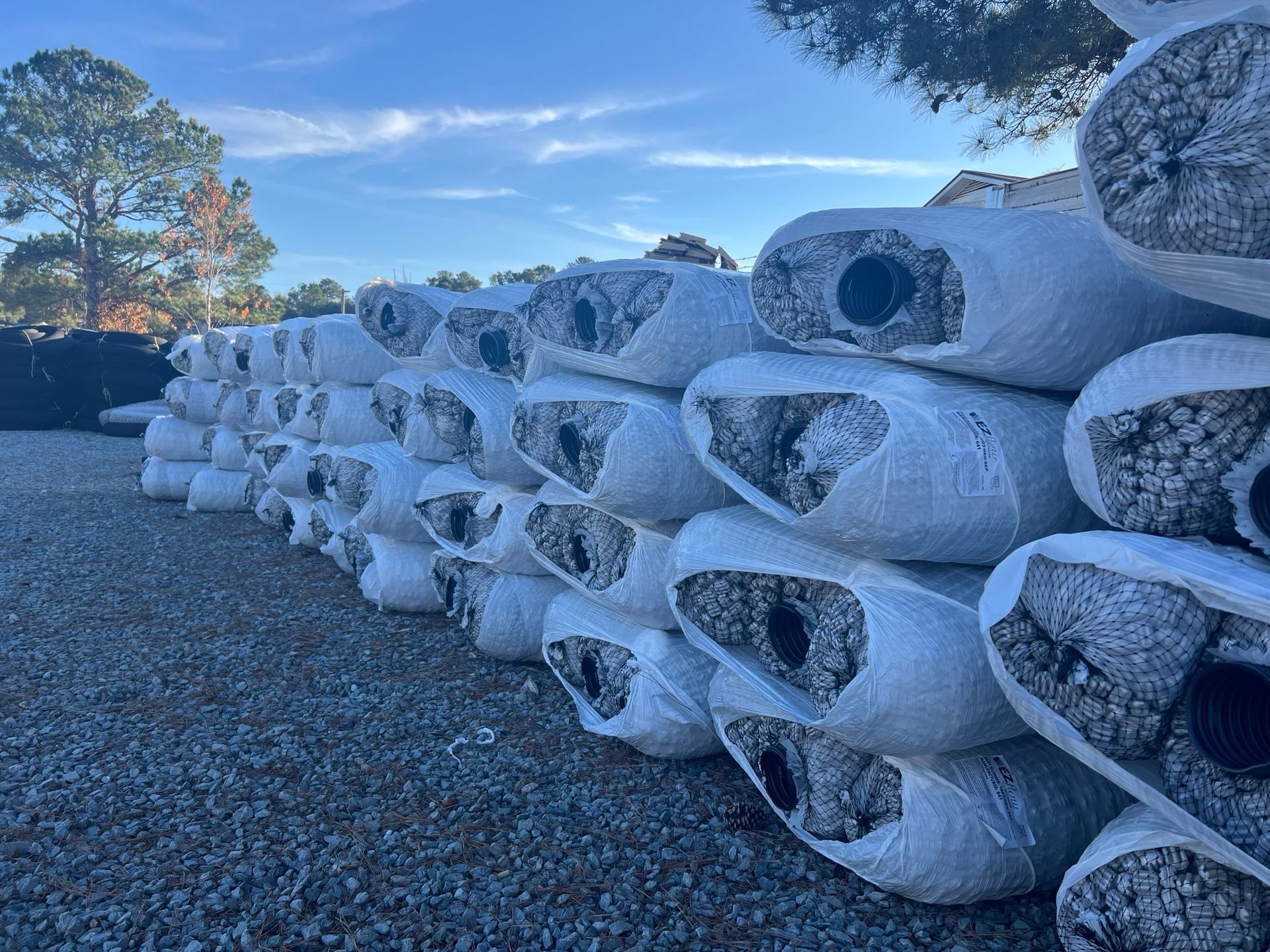 Large rolls of landscape fabric, wrapped in white plastic, stacked outdoors against a blue sky.