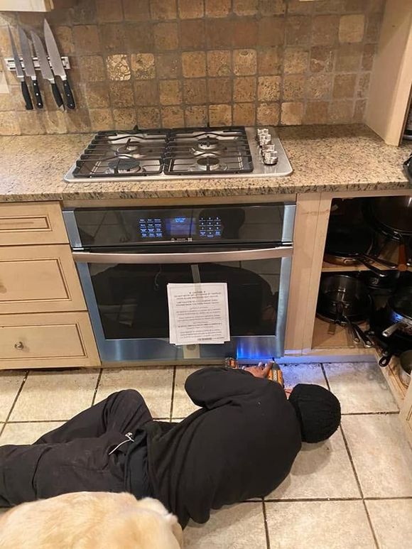 Person in black clothes lying on a kitchen floor near an oven with a blue flame visible below.