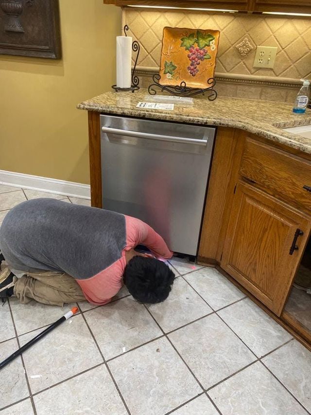 Person crouches on kitchen floor inspecting under a dishwasher near wooden cabinets.
