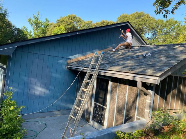 Worker on a roof using a ladder beside a blue house, with debris below.