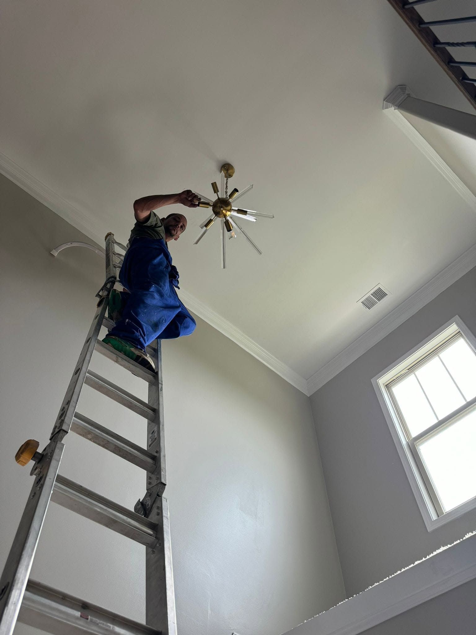 Person on a ladder replacing a ceiling light fixture in a room with white walls and a window.