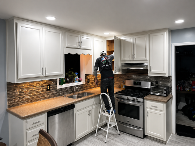 Person on a step stool painting white kitchen cabinets in a bright kitchen.