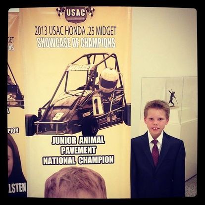 A young boy in a suit and tie stands in front of a usac honda 25 midget showcase of champions poster