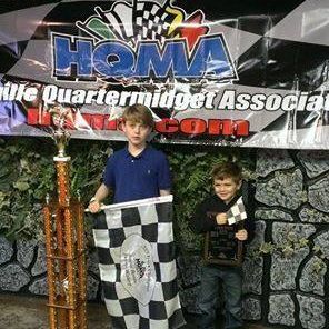 Two young boys are standing next to each other holding a checkered flag and a trophy.