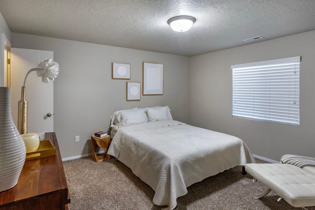 Bedroom with a white bed, nightstand, and window blinds in a neutral-toned room.