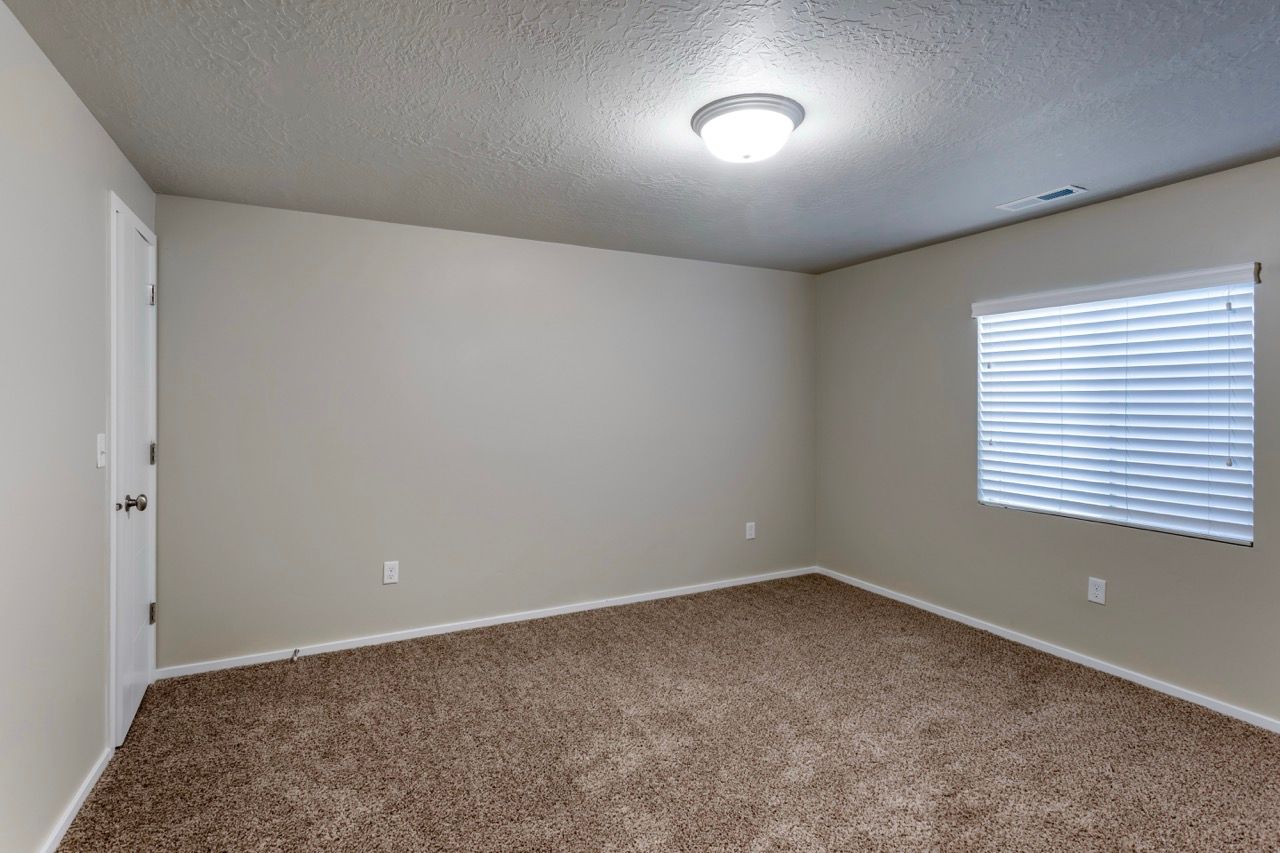 Empty beige-walled room in an apartment with carpet, window blinds, and a ceiling light.