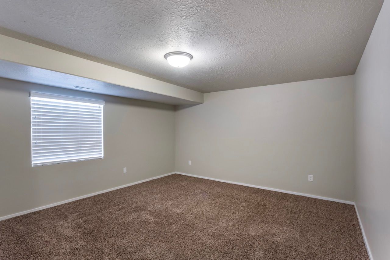 Empty beige apartment room with a window, blinds, and brown carpet.