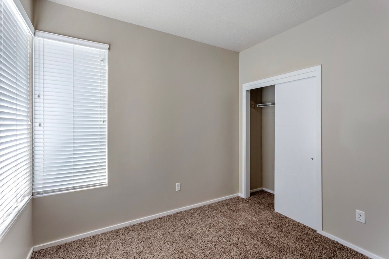 Empty beige apartment bedroom with carpet, large window with blinds, and an open closet.