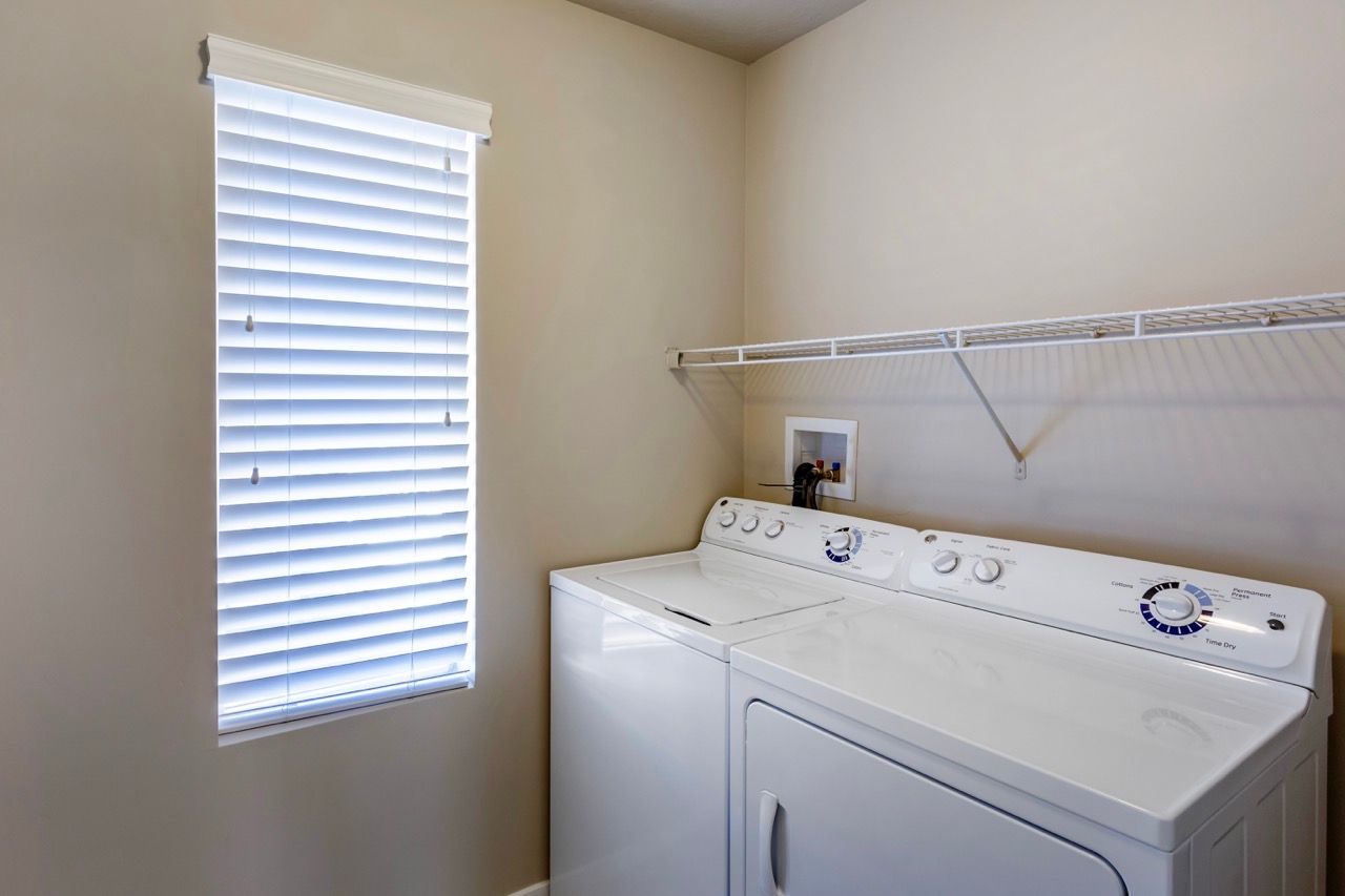 Laundry room with white washer and dryer, window with blinds, and a wire shelf above.