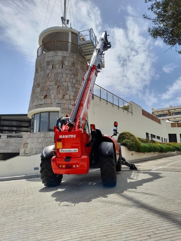 Sollevatore telescopico Manitou rosso proteso verso un edificio in pietra. Giornata luminosa e soleggiata.